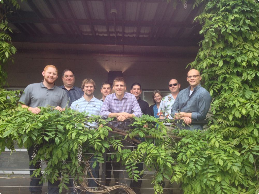 Group of nine people smiling at the camera outside on a platform with vines surrounding them - Applied CAx Siemens software partner USA