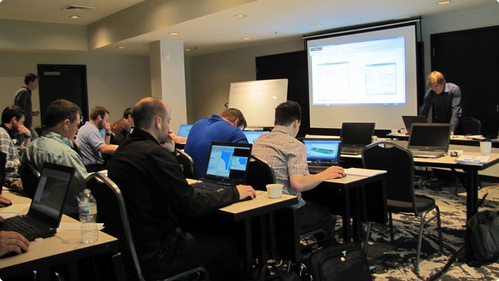 Group of people working on computers in a classroom with a femap tutorial on the board at the front - Applied CAx Siemens software partner USA